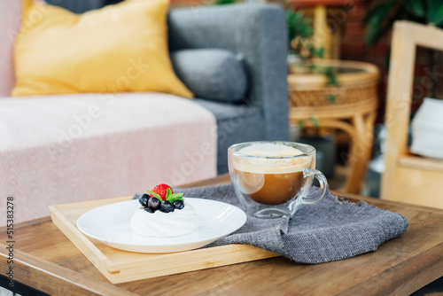 Pavlova dessert mini cake nest and a cup of latte coffee on the wooden tray with gray napkin on the table in cafe. Coffee time. To take a break and relax. Selective focus. Copy space.