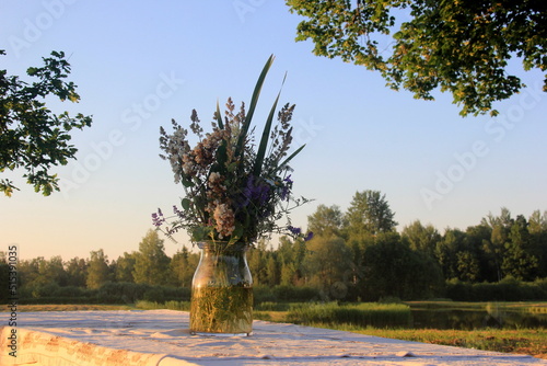 A large wooden table with a white tablecloth and a vase of meadow flowers stands in a green meadow under large trees on the shore of a lake, a Summer Solstice food table in the yellow sunset light