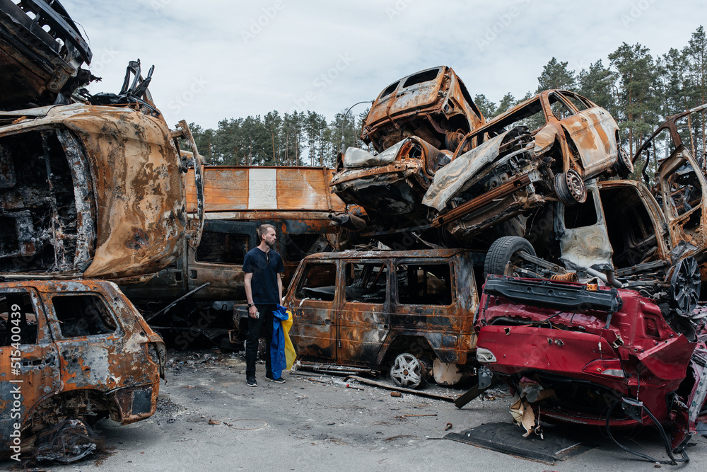 Fototapeta premium Man with Ukrainian flag stands against cars cemetery in Irpin. Consequences of the Russian military invasion in Ukraine.