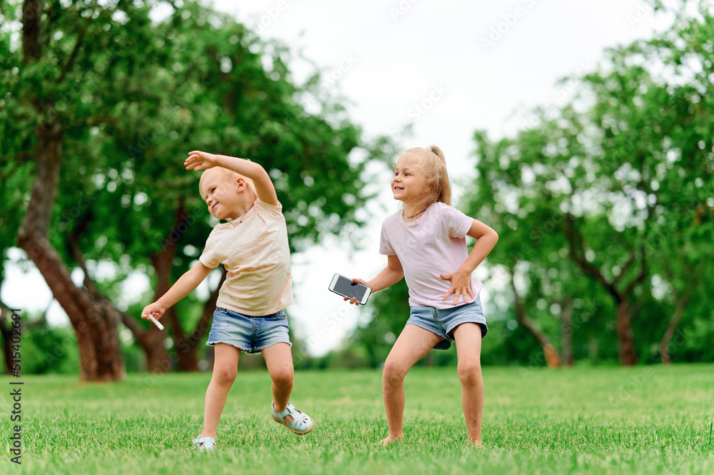 Two little girls sisters dance in the summer in the park on the grass and hold a mobile phone. Family vacation in nature. Happy childhood, healthy offspring Two little girls sisters dance in the summer in the park on the grass and hold a mobile phone. Family vacation in nature. Happy childhood, healthy offspring