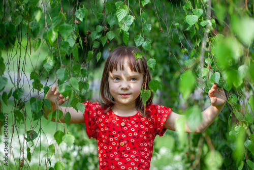 Portrait of a little blonde girl in a in red dress in the foliage of a birch tree in summer day.