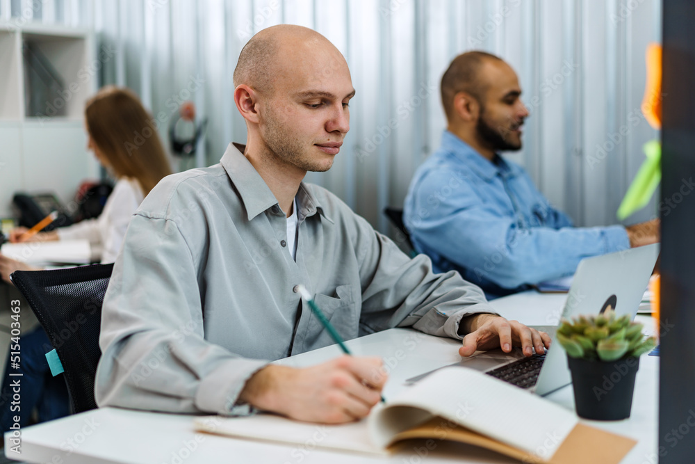Young bald business man sitting at desk in office, working on computer Young bald business man sitting at desk in office, working on computer