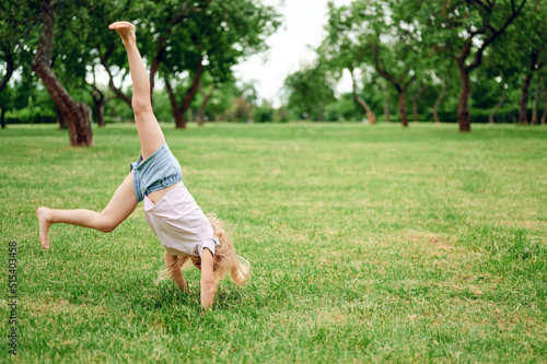 Little blonde girl tumbles on the grass in a summer park. The child is having fun in nature. Happy childhood. Family outdoor recreation with the whole family.