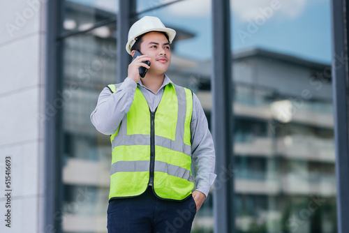Portrait of Asian engineer young man wearing safety vest and helmet standing use smartphone for contact work on building construction site background. Engineering construction worker concept. © Oulaphone