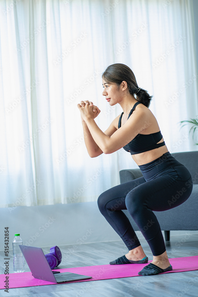 Obraz premium Beautiful Asian woman doing yoga at home A healthy smile stretches over her shoulders at home in the living room. near a large window Full-body portrait, sports, and healthy lifestyle concepts