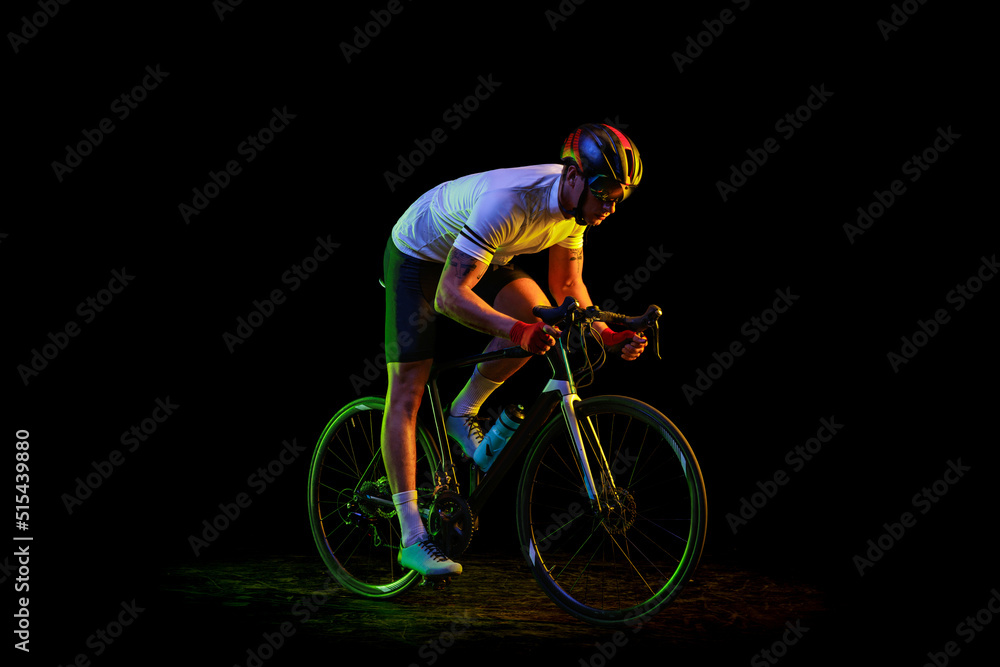 Fototapeta premium Portrait of young male cyclist on bicycle in cycling shorts and protective helmet isolated on dark background in neon. Concept of sport, speed, energy