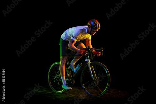 Fototapeta Naklejka Na Ścianę i Meble -  Portrait of young male cyclist on bicycle in cycling shorts and protective helmet isolated on dark background in neon. Concept of sport, speed, energy