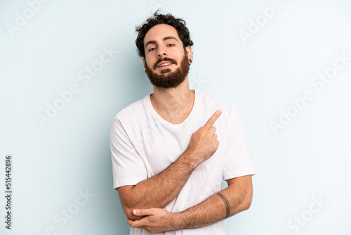 Fototapeta young adult bearded man smiling cheerfully, feeling happy and pointing to the si