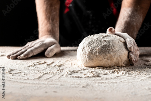 Beautiful and strong men's hands knead the dough from which they will then make bread, pasta or pizza. A cloud of flour flies around like dust. Food concept