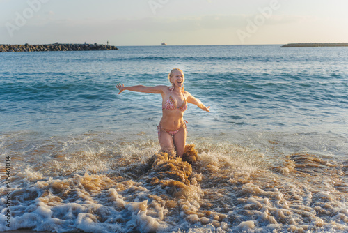 Blond girl is happy by the sea. She opens arms and shouts. Splash of water gives drive. Wavy weather. Human on holidays, rest, tourism, wellnes, happy lifestyle.