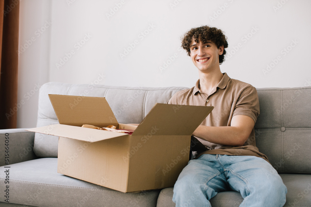 Happy young smiling curly man opening box with ordered goods gifts, presents at home on couch. Online shopper male customer opening online shop parcel. Happy young smiling curly man opening box with ordered goods gifts, presents at home on couch. Online shopper male customer opening online shop parcel.