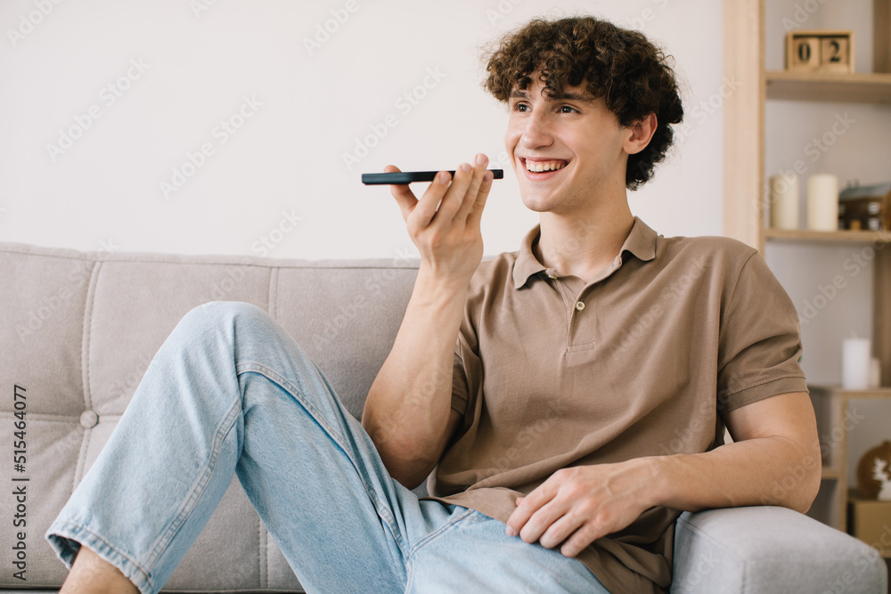 Attractive young male student is recording a voice message on a smartphone while sitting on a sofa in living room Attractive young male student is recording a voice message on a smartphone while sitting on a sofa in living room