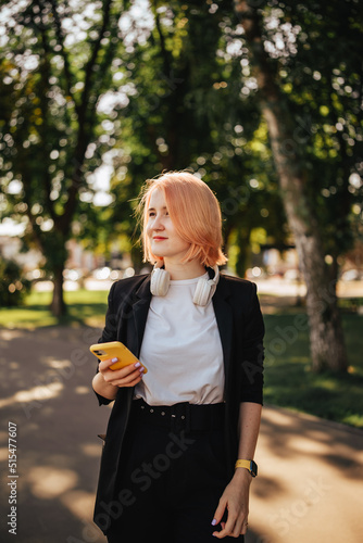 Young blonde woman wearing headphones and holding mobile phone device in the park outdoors in the sunny weather day