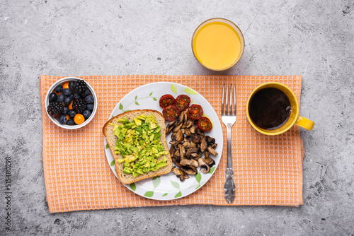 Vegan breakfast setup: avocado toast with fried tomatoes and mushrooms, berries, orange juice, and coffee. Top view