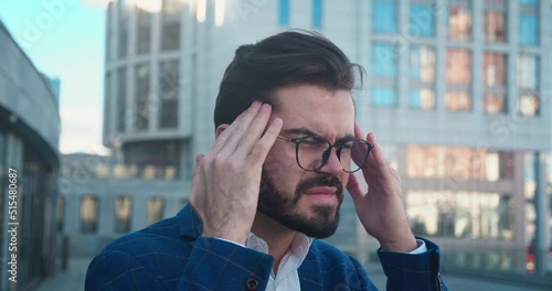 Stressed young man in glasses suffering from muscles tension, having painful head feelings due to. Tired employee overwhelmed with tasks near bussinescenter standing in outdoor. photo