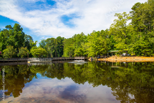 a gorgeous summer landscape in the park with a lake and a water fountain and a brown wooden bridge surrounded by lush green trees, grass and plants with blue sky and clouds at Dupree park in Woodstock photo