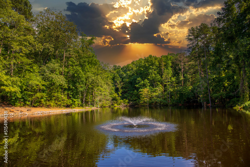 Canvas Print a still lake with a water fountain in the center surrounded by lush green trees