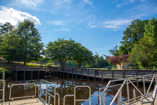 a gorgeous summer landscape in the park with a lake and a brown wooden bridge surrounded by lush green trees, grass and plants with blue sky and clouds at Dupree Park in Woodstock Georgia USA photo