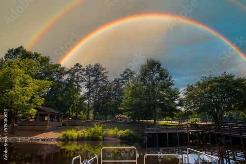 a gorgeous summer landscape in the park with a lake and a brown wooden bridge surrounded by lush green trees, grass and plants with blue sky, clouds and rainbow at Dupree Park in Woodstock Georgia USA photo