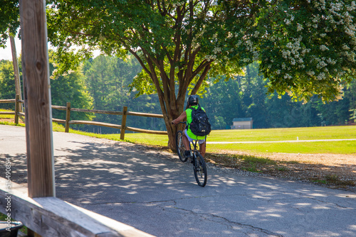 a man wearing a yellow shirt and a black helmet and backpack riding a bike in the park with a gorgeous summer landscape surrounded by lush green trees, grass and plants at Dupree Park in Woodstock photo