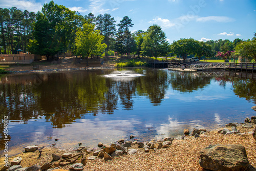 a gorgeous summer landscape in the park with a lake and a water fountain and a brown wooden bridge surrounded by lush green trees, grass and plants with blue sky and clouds at Dupree park in Woodstock