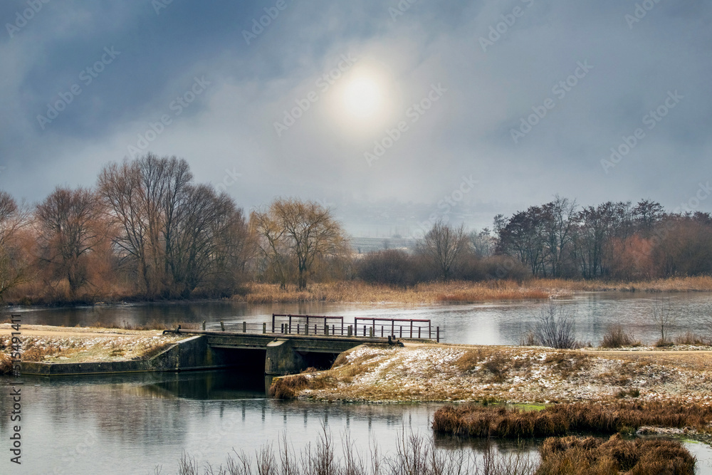 Winter landscape with trees near the river and cloudy sky with dim sun, bridge over the river. The beginning of winter, the first snow