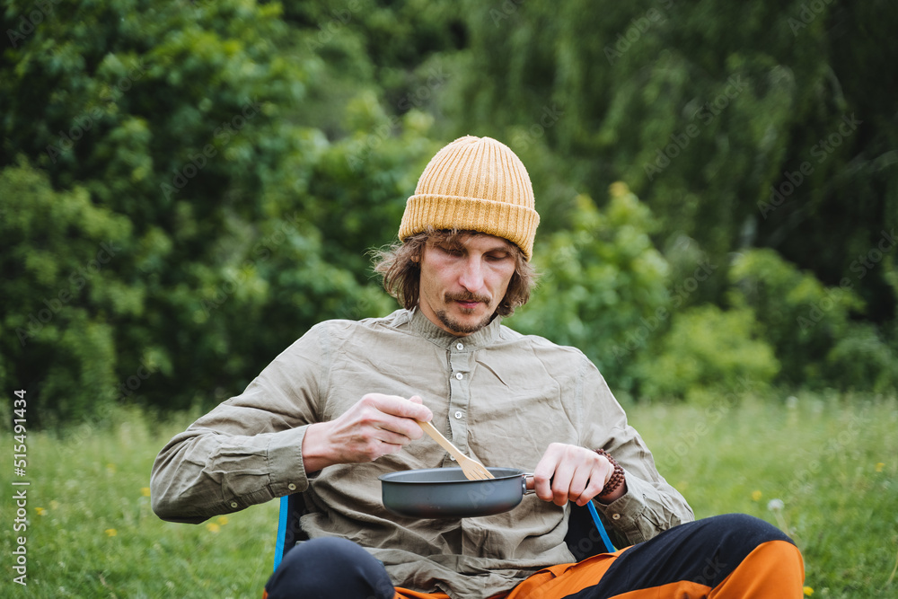 Fototapeta premium A person on a hike eats food from a frying pan, tourist food in nature, a guy on a hike has breakfast with scrambled eggs, a summer vacation in nature camping