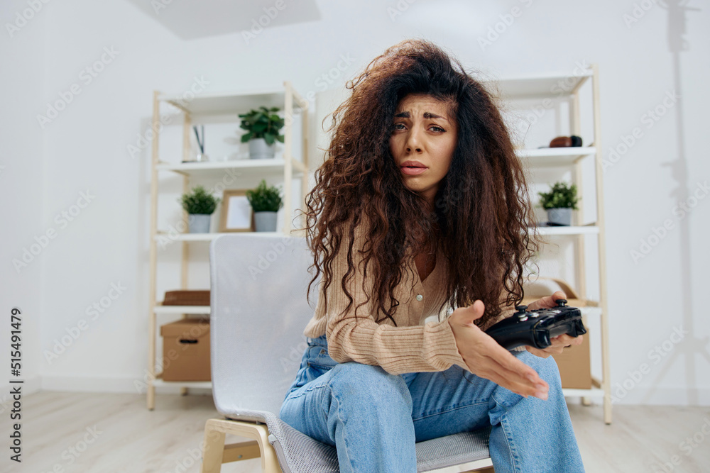 Upset sad frustrated tanned pretty curly Latin lady lose in game with joystick gamepad sit on chair in home modern interior look at camera. Copy space Mockup Banner. Concept gamer, emotions Upset sad frustrated tanned pretty curly Latin lady lose in game with joystick gamepad sit on chair in home modern interior look at camera. Copy space Mockup Banner. Concept gamer, emotions