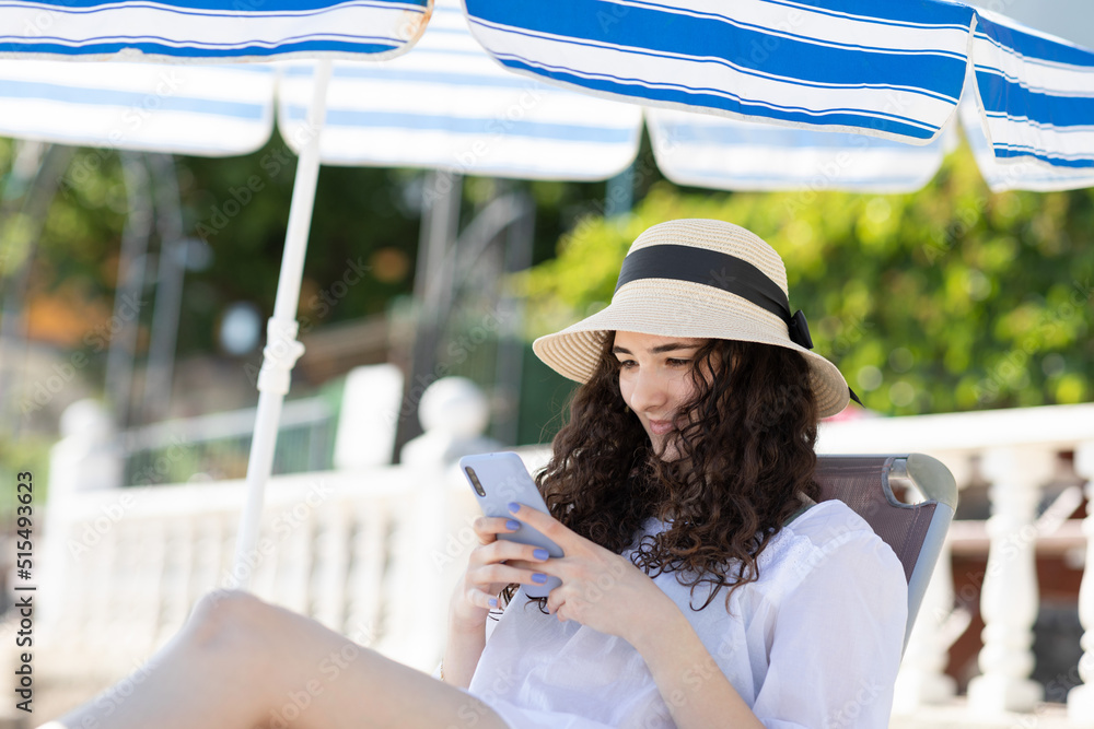 Young smiling teenager girl texting on the mobile phone sitting on the sunbed. Young woman using smartphone. Young smiling teenager girl texting on the mobile phone sitting on the sunbed. Young woman using smartphone.