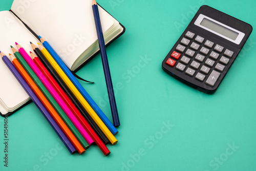 Several colored pencils on top of a notebook and a black calculator on a green table. Back to school.