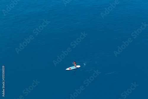 Top view of a group of surfers in the water. Guys and girls are waiting for a wave. Some just swim. Sandy bottom. The rays of the sun on the water. Small waves. Colored boards