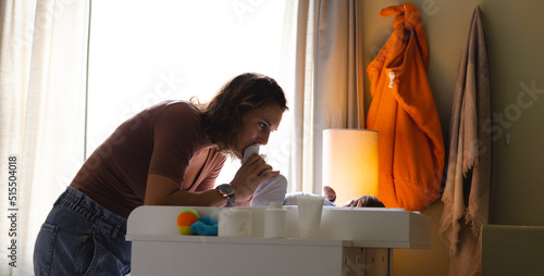 Caucasian mid adult mother playing with newborn baby on nappy changing table by window at home photo