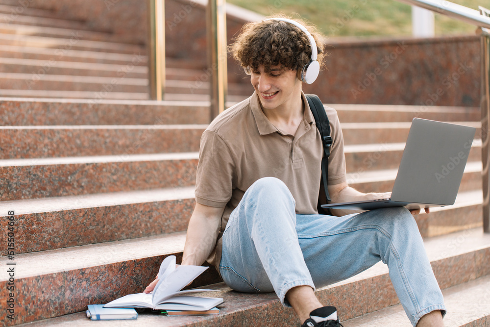 Concept of e-learning, distance study or remote learning concept, Young happy school guy, college or university student with backpack and headphones using laptop sitting on steps in university