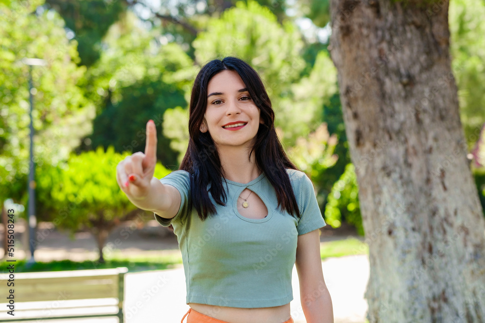 Cheerful brunette woman wearing tee standing on city park, outdoors showing and pointing up with fingers number one while smiling confident and happy. Selective focus on her face. Cheerful brunette woman wearing tee standing on city park, outdoors showing and pointing up with fingers number one while smiling confident and happy. Selective focus on her face.