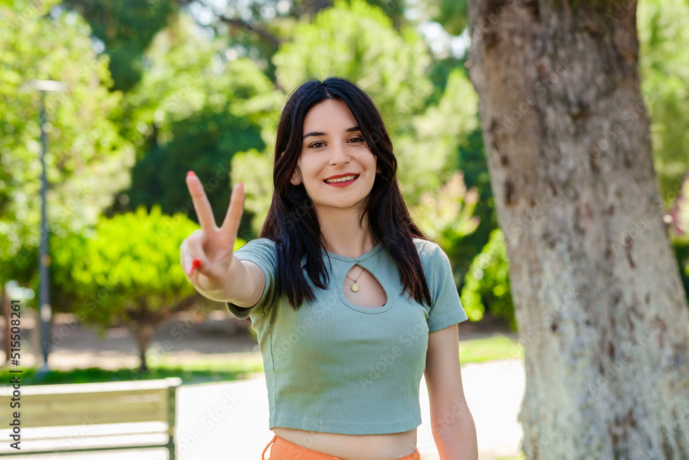 Happy brunette woman wearing turquoise tee on city park, outdoors showing and pointing up with fingers number two while smiling confident and happy. Selective focus on her face. Happy brunette woman wearing turquoise tee on city park, outdoors showing and pointing up with fingers number two while smiling confident and happy. Selective focus on her face.