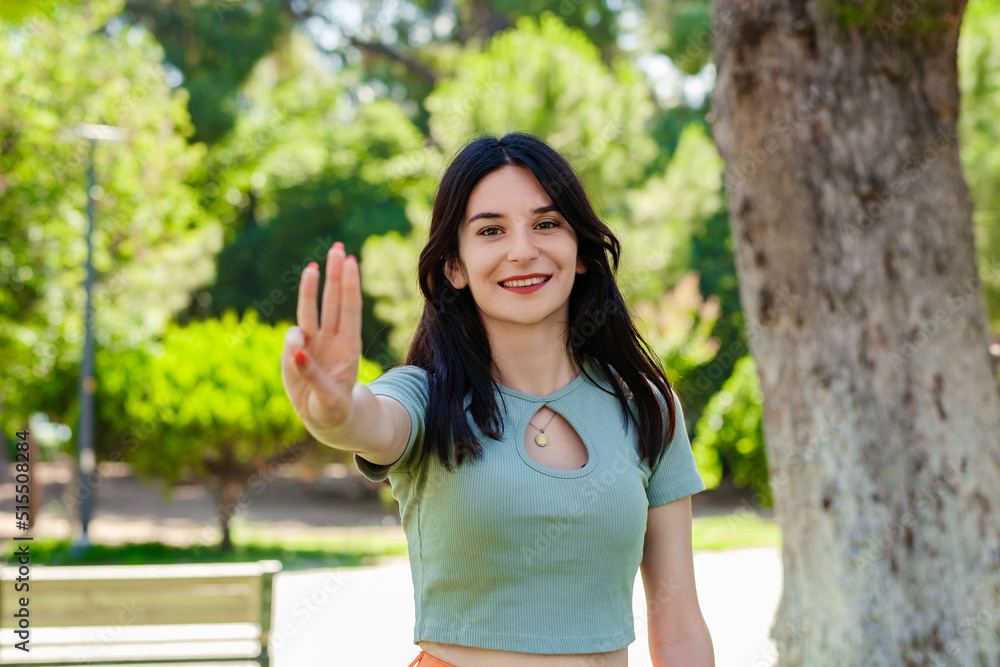 Happy woman wearing tee standing on city park, outdoors showing and pointing up with fingers number three while smiling confident and happy. Selective focus on her face. Happy woman wearing tee standing on city park, outdoors showing and pointing up with fingers number three while smiling confident and happy. Selective focus on her face.