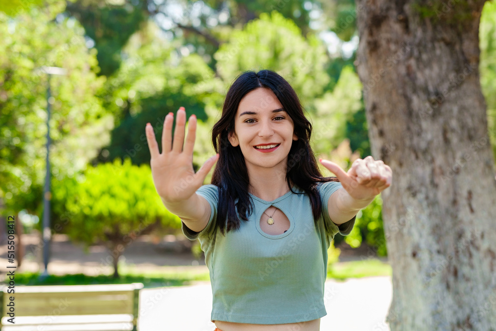 Happy woman wearing tee standing on city park, outdoors showing and pointing up with fingers number six while smiling confident and happy. Selective focus on her face. Happy woman wearing tee standing on city park, outdoors showing and pointing up with fingers number six while smiling confident and happy. Selective focus on her face.