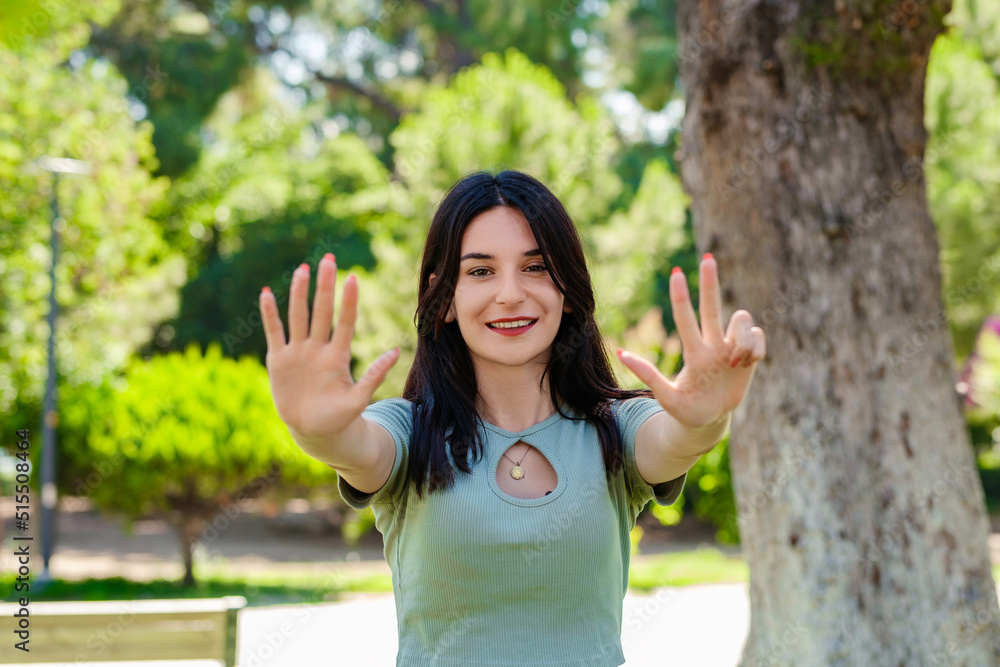 Cheerful brunette woman wearing tee standing on city park, outdoors showing and pointing up with fingers number eight while smiling confident and happy. Selective focus on her face. Cheerful brunette woman wearing tee standing on city park, outdoors showing and pointing up with fingers number eight while smiling confident and happy. Selective focus on her face.