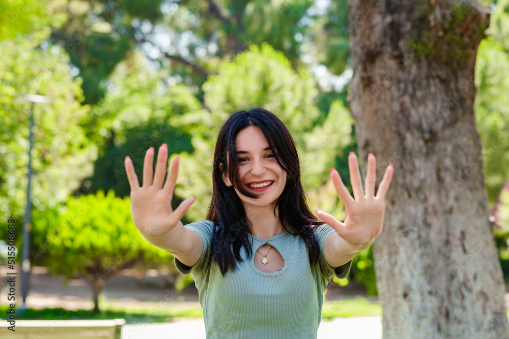 Happy brunette woman wearing turquoise tee on city park, outdoors showing and pointing up with fingers number ten while smiling confident and happy. Selective focus on her face. Happy brunette woman wearing turquoise tee on city park, outdoors showing and pointing up with fingers number ten while smiling confident and happy. Selective focus on her face.