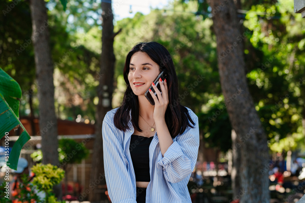 Young brunette woman wearing blue striped shirt on city park, outdoors talking on mobile phone with friends or boyfriend with smiles. Young brunette woman wearing blue striped shirt on city park, outdoors talking on mobile phone with friends or boyfriend with smiles.