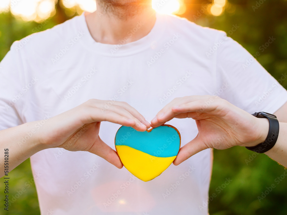 Man in a white T-shirt holds in his hands a gingerbread in the shape of a heart in the colors of the flag of Ukraine yellow and light blue. Assistance, help and solidarity with Ukraine. High quality