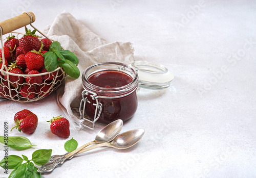 Strawberry jam with basil in the glass jar and a basket with berries at white table. Copy space