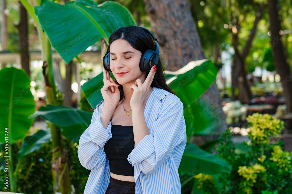 Brunette millennial woman wearing striped shirt standing on city park, outdoors listening music in headphones and eyes closed. Enjoy good quality sound, stress free concept. Brunette millennial woman wearing striped shirt standing on city park, outdoors listening music in headphones and eyes closed. Enjoy good quality sound, stress free concept.