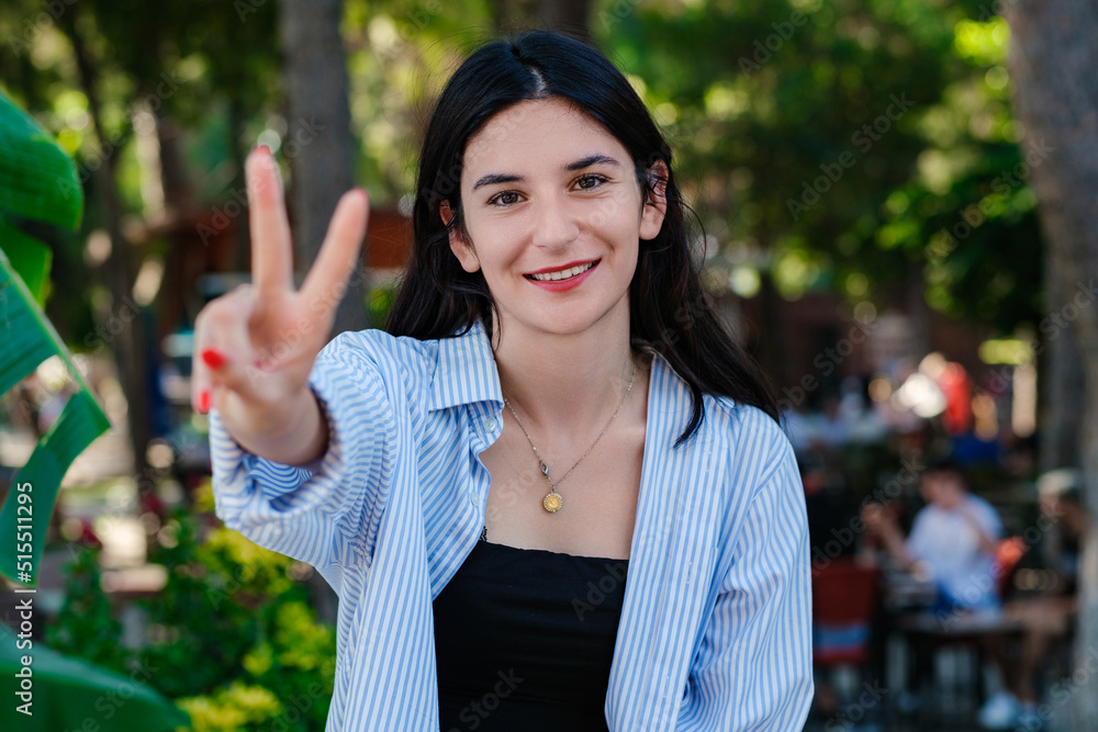 Young brunette girl smiling happy wearing blue striped shirt on city park, outdoors showing and pointing up with fingers number two while smiling confident and happy. Selective focus on her face. Young brunette girl smiling happy wearing blue striped shirt on city park, outdoors showing and pointing up with fingers number two while smiling confident and happy. Selective focus on her face.