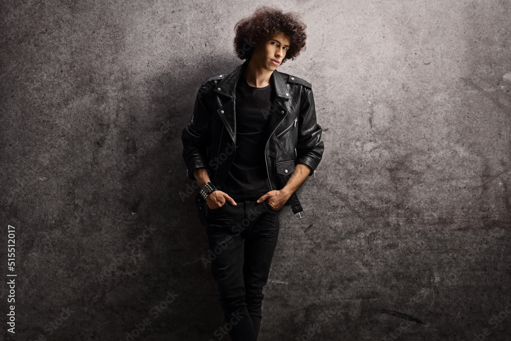 Young man in a leather jacket and curly hair leaning on a gray rugged wall