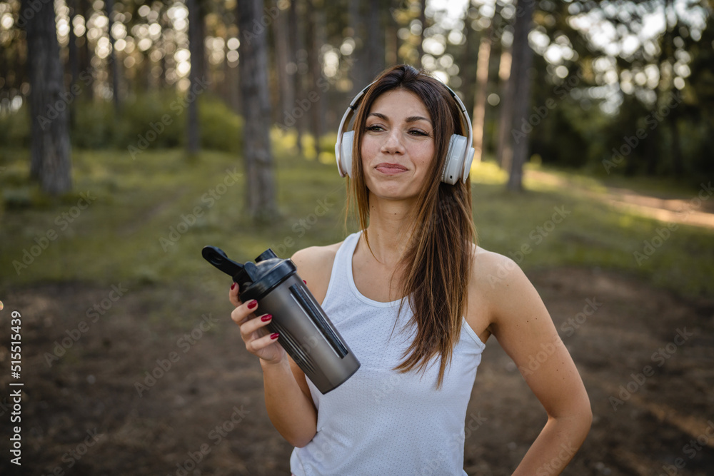 One woman happy caucasian female standing in the forest or park in woods with supplement shaker and headphones during training in nature taking a brake water drink and hydration real people copy space One woman happy caucasian female standing in the forest or park in woods with supplement shaker and headphones during training in nature taking a brake water drink and hydration real people copy space