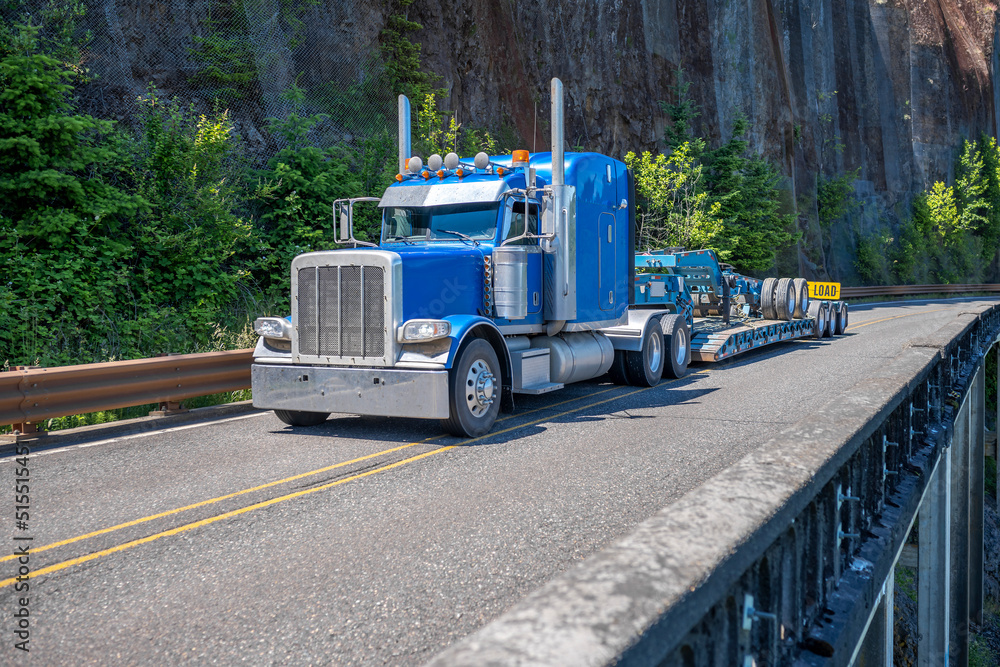 Blue classic big rig semi truck with chrome accessories and extended cab transporting step down semi trailer extender fastened on step down semi trailer running on the mountain road