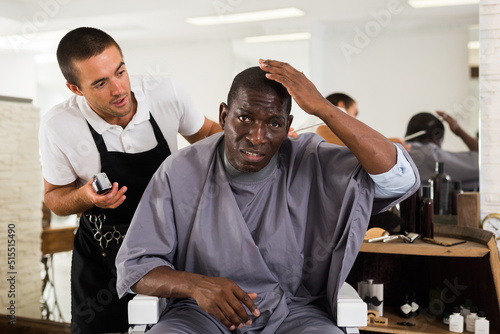 Portrait of shocked African American sitting in barber chair with confused young hairdresser behind him