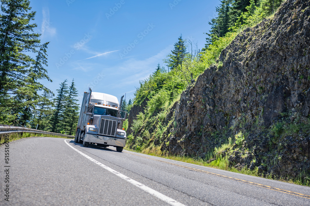 Heavy duty big rig semi truck with dry van semi trailer running on the turning winding mountain highway road with rocks and trees on the side