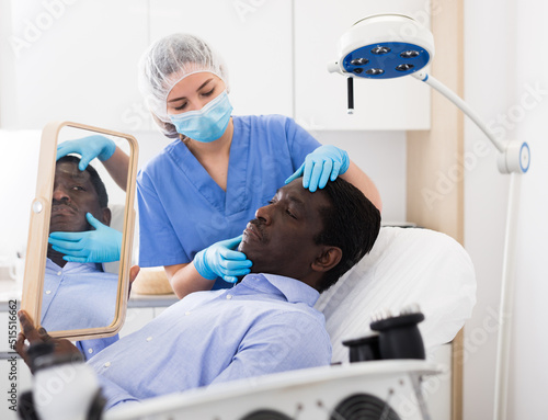 Professional cosmetologist examining male client face before procedure in esthetic clinic. Focused man looking at mirror